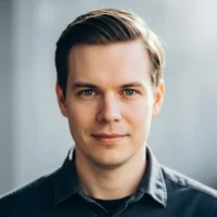 Professional headshot of a smiling man with neatly styled brown hair, blue-green eyes, and a dark shirt.
